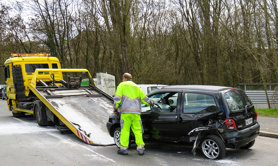 Photo of yellow flatbed towing truck about to tow damaged black car with employee standing in the side moving the steering wheel. Photo of yellow flatbed towing truck about to tow damaged black car with employee standing in the side moving the steering wheel.